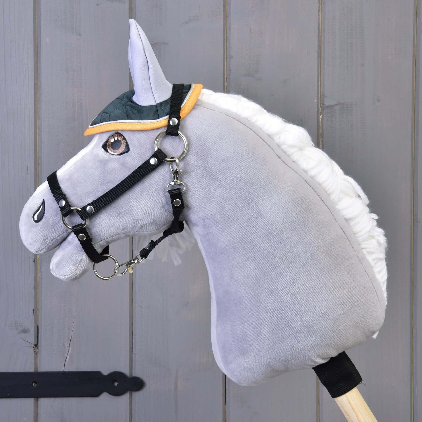 Čabraka Thermo Sport od Hobby Horse LarDen v zelenej a žltej farbe na sivom plyšovom Hobby Horse s bielou hrivou a čiernou uzdečkou, nasadenom na drevenej tyči pred sivou drevenou stenou.